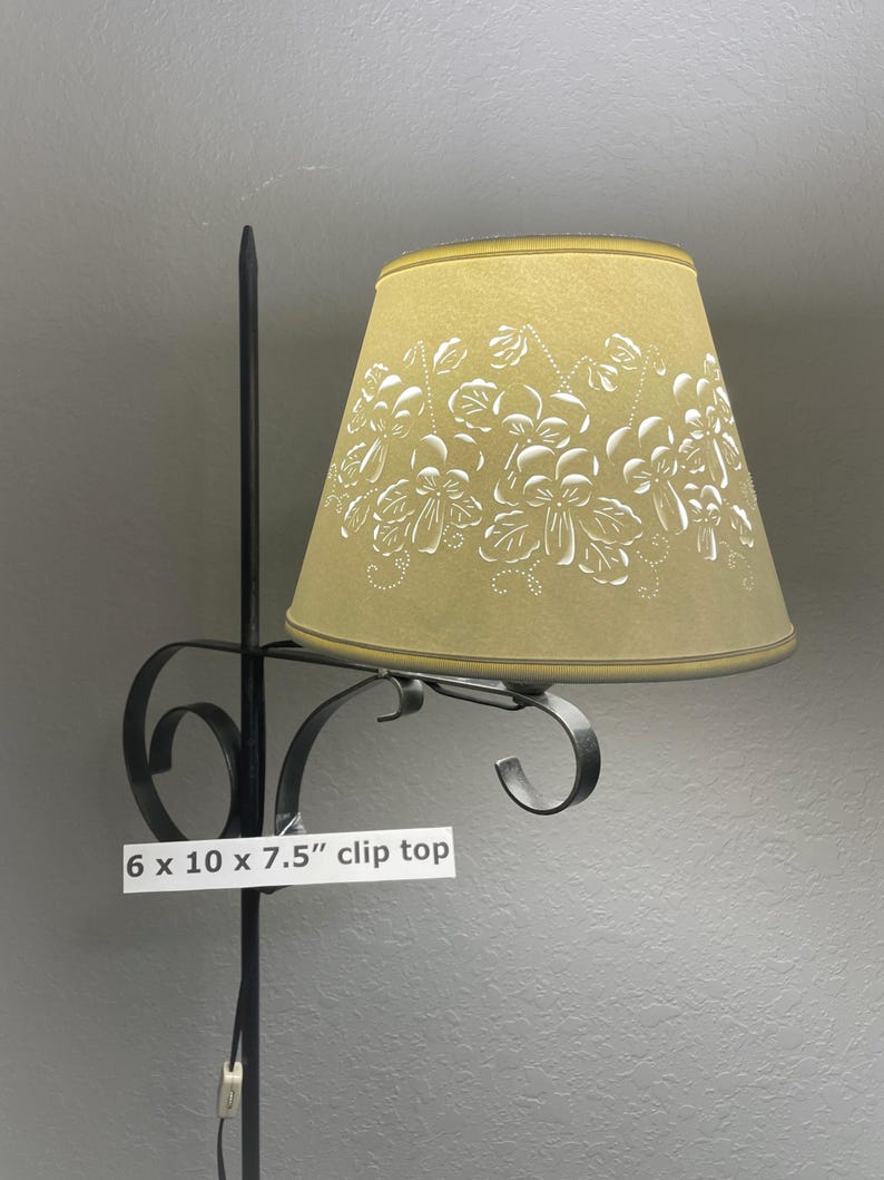 May include: A clip-top lampshade with a floral design. The shade is 6 inches wide, 10 inches long, and 7.5 inches tall. The shade is a light yellow color with white floral patterns. The lamp has a black metal frame.