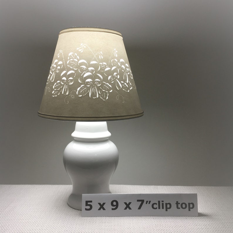 May include: A white ceramic table lamp with a beige lampshade. The lampshade features a floral design. The lamp is 5 inches wide, 9 inches long, and 7 inches tall. The lamp has a clip top.