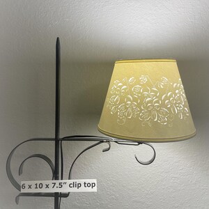 May include: A clip-top lampshade with a floral design. The lampshade is 7.5 inches tall, 10 inches wide, and 6 inches deep. The lampshade is a light yellow color with white floral patterns. The metal frame is black.