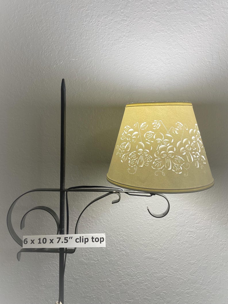 May include: A clip-top lampshade with a floral design. The lampshade is 7.5 inches tall, 10 inches wide, and 6 inches deep. The lampshade is a light yellow color with white floral patterns. The metal frame is black.
