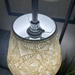 May include: A close-up of a lamp with a beige lampshade featuring a leaf pattern. The lamp's metal frame is black, and the light bulb is visible inside the shade. The lamp is turned on, illuminating the leaf design.