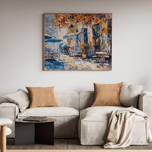 May include: A white sectional sofa with two brown throw pillows and a white throw blanket. The sofa is in front of a white wall with a painting of a cafe scene above it. The painting is framed in a light wood frame.
