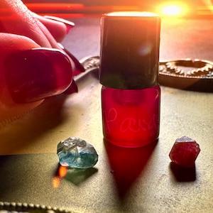 May include: A small glass bottle with a black cap filled with a red liquid. The bottle is sitting on a gold surface with two small gemstones, one green and one red.