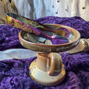 May include: A ceramic incense burner with a handle, filled with grey ash and a pair of iridescent metal incense tongs. The burner has a brown and beige glaze with a decorative design.