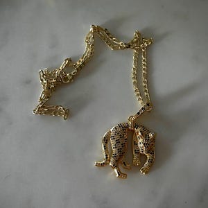 以下が含まれることがあります： 黒いアクセントが付いた、金色のスタイリッシュなヒョウのペンダントが付いたネックレス。チェーンはクラシックなリンクデザインです。ネックレスは明るい色の表面に表示され、ジュエリーの詳細が強調されています。