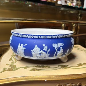 Może przedstawiać: Okrągła, niebieska ceramiczna miska z białymi figurami w reliefie przedstawiającymi klasyczne sceny na zewnątrz. Miska ma białe wnętrze, ozdobną krawędź z motywem liści wzdłuż górnego brzegu i stoi na trzech małych, zakrzywionych białych nóżkach.