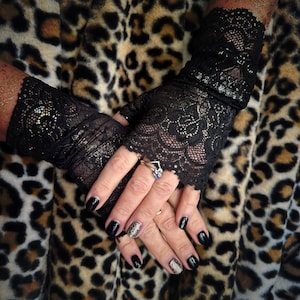May include: A pair of black lace fingerless gloves with a sparkly finish. The gloves are worn on a person's hands with black nail polish and glitter nail polish.