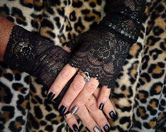 May include: A pair of black lace fingerless gloves with a sparkly finish. The gloves are worn on a person's hands with black nail polish and glitter nail polish.
