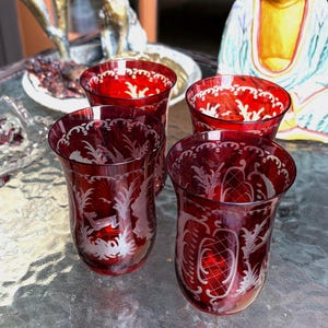 Könnte beinhalten: Ein Set aus vier tiefroten Glasbechern mit detaillierten, weiß geätzten Motiven von Hirschen, Blattwerk und geometrischen Mustern. Jedes Glas hat einen ausgestellten Rand und einen abgerundeten Boden und besticht durch eine durchscheinende rubinrote Farbe.