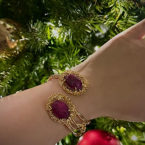May include: A gold-toned cuff bracelet featuring two oval, deep red gemstones. The gemstones are set within ornate, textured gold frames. The bracelet's design includes delicate wirework and is displayed against a backdrop of Christmas tree greenery and ornaments.