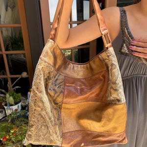 May include: A hobo-style shoulder bag featuring a patchwork design of metallic bronze leather and textured snakeskin-patterned panels. The bag includes a single wide shoulder strap attached with rectangular metal hardware and a spacious main compartment.