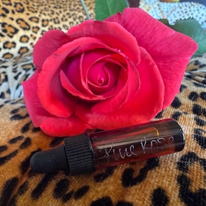 May include: A vibrant red rose with velvety petals next to a small brown glass bottle. The bottle has a black dropper and the words "Blue Rose" are handwritten on the side. The background has a leopard print pattern.
