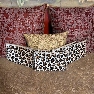 May include: Three rectangular pouches featuring a leopard print pattern on a white and tan fur-like texture. Each pouch has a silver-toned zipper closure along the top edge and is constructed with visible stitching along the sides and bottom.
