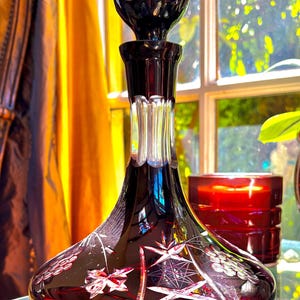 May include: A ruby red glass decanter with a round, bulbous base and a tall, slender neck. The decanter features intricate cut glass designs, including stars and floral patterns. The stopper is a dark, round shape. The decanter is displayed on a glass surface.