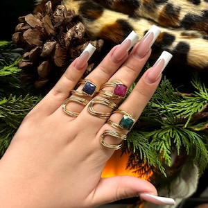 May include: A hand displays three gold-toned rings, each featuring a square gemstone in blue, red, and green. The rings have a unique coiled design. The background includes a leopard print pattern and pine needles.
