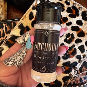 Puede incluir: Un frasco de plástico transparente con tapa negra que contiene polvo corporal. La etiqueta negra presenta texto blanco que dice PATCHOULI DUST OF THE JOURNEY BODY POWDER ATHENA'S LAIR, enmarcado por líneas geométricas. El frasco contiene polvo claro.