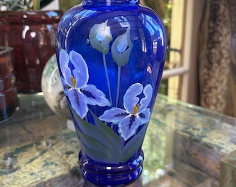 Puede incluir: Un florero de vidrio azul cobalto con un diseño pintado a mano de flores de iris de color morado claro con hojas y tallos verdes. El florero tiene un cuerpo redondeado, un cuello estrecho y un borde ensanchado, con un acabado translúcido.
