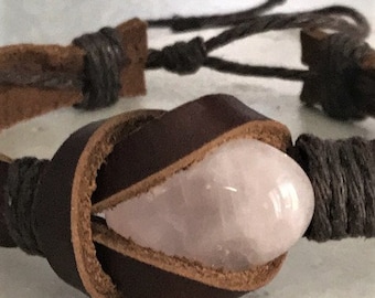 May include: A close-up of a brown leather bracelet featuring a light pink rose quartz stone. The stone is encased in a dark brown leather band. The bracelet has a braided cord closure.