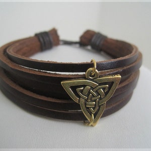 May include: A brown leather bracelet with multiple strands and a gold-colored Celtic knot charm. The bracelet has a simple, rustic design, suitable for everyday wear. The charm is a detailed, triangular shape.
