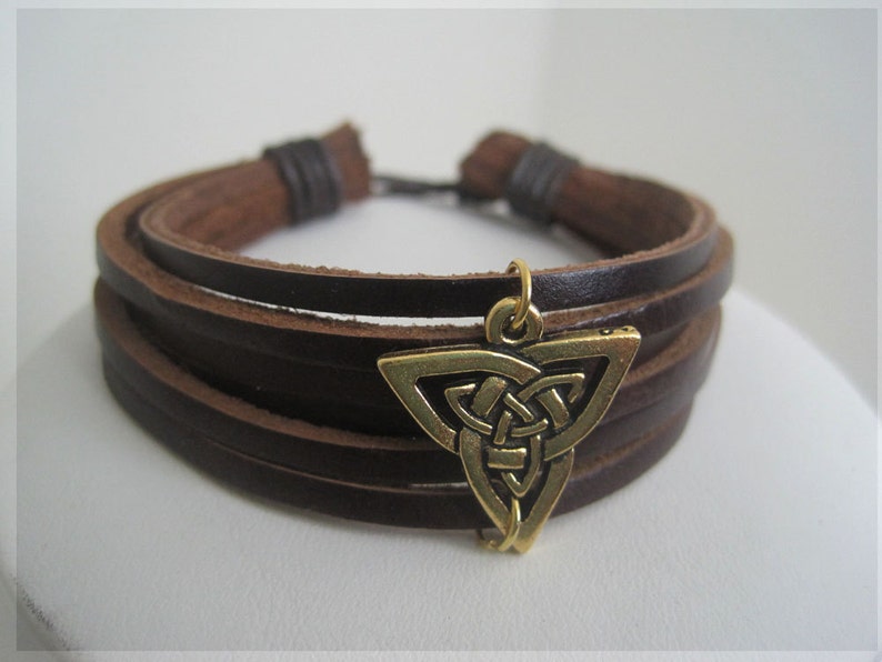 May include: A brown leather bracelet with multiple strands and a gold-colored Celtic knot charm. The bracelet has a simple, rustic design, suitable for everyday wear. The charm is a detailed, triangular shape.