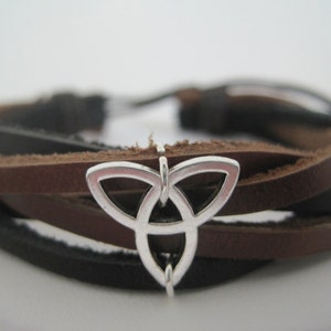 May include: A brown leather bracelet with a silver Celtic knot charm. The bracelet is made of multiple strands of leather, woven together.