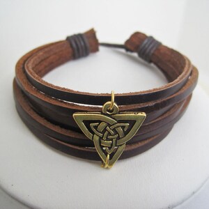 May include: A brown leather bracelet with multiple strands and a bronze-colored Celtic knot charm. The bracelet is adjustable and features a simple design, suitable for everyday wear. The charm is a triangle shape with an intricate knot design.