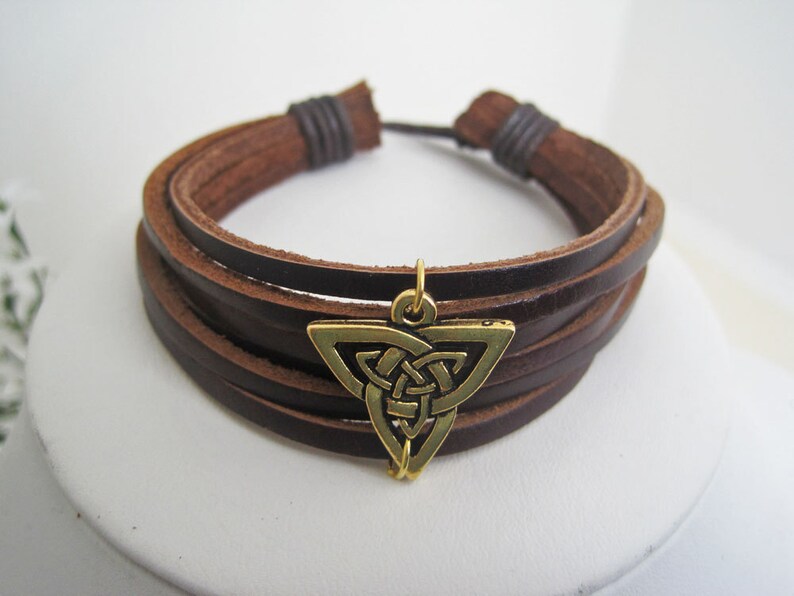 May include: A brown leather bracelet with multiple strands and a bronze-colored Celtic knot charm. The bracelet is adjustable and features a simple design, suitable for everyday wear. The charm is a triangle shape with an intricate knot design.