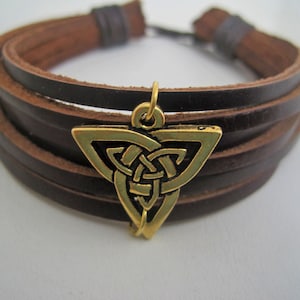 May include: Brown leather bracelet with multiple strands and a gold-colored Celtic knot charm. The bracelet has a clasp closure and is designed for a comfortable fit. The charm adds a touch of elegance to the rustic design.