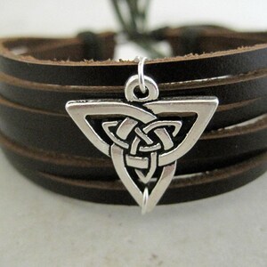 May include: A dark brown leather bracelet with a silver-colored Celtic knot charm. The bracelet features multiple leather bands and a clasp. The charm is a triangular shape with an intricate knot design.