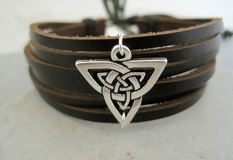 May include: A dark brown leather bracelet with a silver-colored Celtic knot charm. The bracelet features multiple leather bands and a clasp. The charm is a triangular shape with an intricate knot design.
