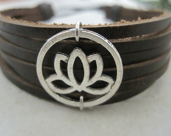 May include: A dark brown leather bracelet with multiple bands. A silver-colored circular charm with a lotus flower design is attached to the bracelet. The bracelet is a close-up shot.