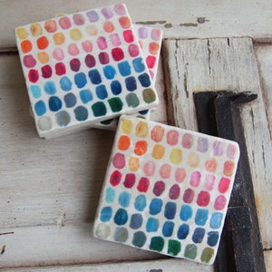 May include: Set of four square coasters with a colorful paint dot design. The coasters are made of a white material and feature a gradient of colors from yellow to blue.