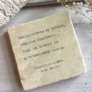 May include: A square, off-white coaster with a quote from Charlotte's Web by E.B. White. The quote, in black typewriter font, reads: "You have been my friend, replied Charlotte. That in itself is a tremendous thing."