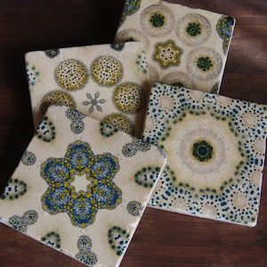 May include: A set of four square ceramic coasters with intricate, colorful patterns. The designs feature yellow, blue, and green circular and floral motifs on a cream-colored background. The coasters are arranged on a dark wooden surface.