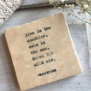 May include: A beige stone coaster with a quote from Ralph Waldo Emerson: "live in the sunshine. swim in the sea. drink the wild air." The quote is printed in black typewriter font.