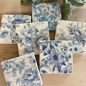 May include: Six square ceramic coasters with a blue and white floral pattern. The coasters are made of marble and have a distressed look.