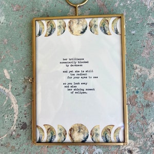 May include: A gold-framed print with a white background and a poem about the moon's brilliance. The poem reads: "her brilliance momentarily blocked by darkness and yet she is still too radiant for your eyes to see so you look away and miss her shining moment of eclipse."