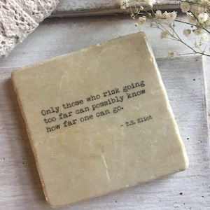May include: A beige square coaster with a quote by T.S. Eliot: "Only those who risk going too far can possibly know how far one can go." The quote is printed in black typewriter font.