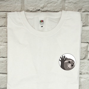 May include: White t-shirt with a small, round, black and white graphic of an otter waving. The otter is looking to the right and has a surprised expression.