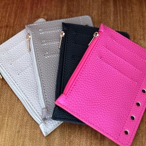 May include: Four small wallets in silver, gray, black, and hot pink. Each wallet has a zipper closure with a gold-colored zipper pull. The hot pink wallet has several holes along the edge, and the wallets are made of textured material.