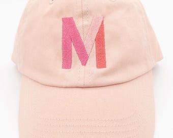 Puede incluir: Gorra de béisbol rosa claro con visera curva. La gorra presenta una gran letra "M" bordada en tonos rosa y coral. El sombrero está hecho de una tela suave y texturizada y tiene un diseño clásico de seis paneles.