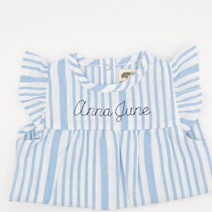 Puede incluir: Un vestido de bebé a rayas azules y blancas con cuello y mangas con volantes. El vestido tiene el nombre "Anna June" bordado en la parte delantera.
