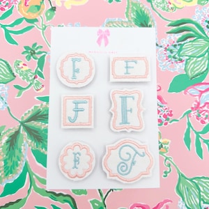May include: A set of six white fabric patches featuring the letter F embroidered in light blue, each bordered with a soft pink stitched edge. The patches are arranged on a white display card with a pink bow icon and the text Magnolia Hall.