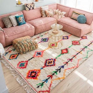 Könnte beinhalten: Ein Wohnzimmer mit einem großen, rosa Ecksofa und einem cremefarbenen Teppich mit farbenfrohem geometrischem Muster. Der Teppich hat einen Fransenrand. Dekorative Kissen und kleine goldene Beistelltische ergänzen die Einrichtung.