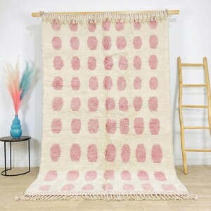 May include: A cream-colored rug with a repeating pattern of pink oval shapes. The rug has a fringe along the bottom edge and is displayed on a wooden rod. The rug is a decorative home decor item.