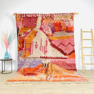 Op de afbeelding: Een grote, kleurrijke Marokkaanse vloerkleed met een geometrisch ontwerp. Het vloerkleed heeft een mix van roze, oranje, rode en paarse tinten. Het wordt getoond op een houten staaf, met een crèmekleurige franje langs de onderrand.