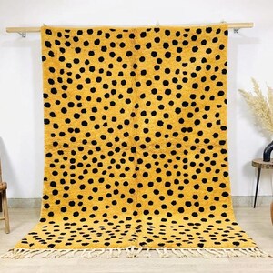 Puede incluir: Una alfombra rectangular con un fondo amarillo mostaza vibrante cubierto por un patrón repetitivo de puntos negros irregulares. La alfombra tiene un pelo grueso y texturizado y está rematada con largos flecos de color crema en el borde.