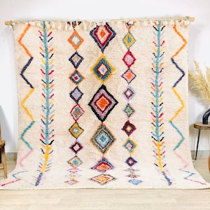 May include: A large, cream-coloured rug with a geometric pattern. The design features colourful diamond shapes in orange, yellow, blue, and pink, arranged in vertical rows. The rug has a textured appearance and is displayed against a white wall.