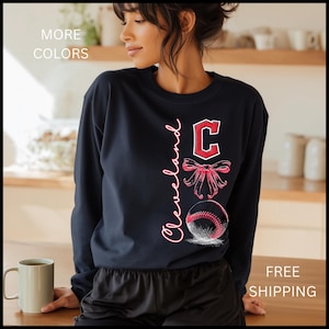 May include: Navy blue sweatshirt with the word "Cleveland" in pink script, a red "C" logo, a pink bow, and a baseball graphic. The text "MORE COLORS" is in the upper left corner.