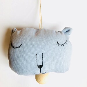 May include: A light blue fabric baby rattle with a wooden ring and a wooden nose. The rattle has embroidered eyes and a stitched mouth.